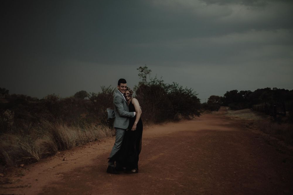 An elegant couple embracing on a desert path with dramatic storm clouds overhead.