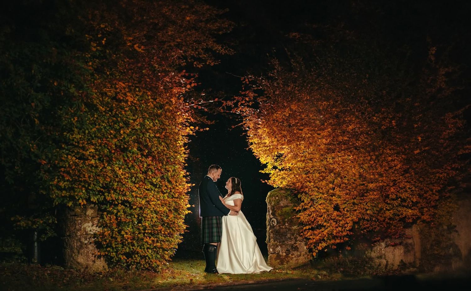 Bride and groom share a romantic moment under autumn foliage arch at a castle venue at night.