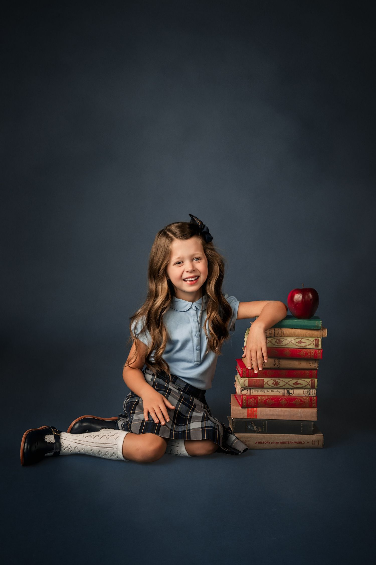 A smiling young girl in school uniform sits beside a stack of vintage books topped with a red apple on a dark studio background.