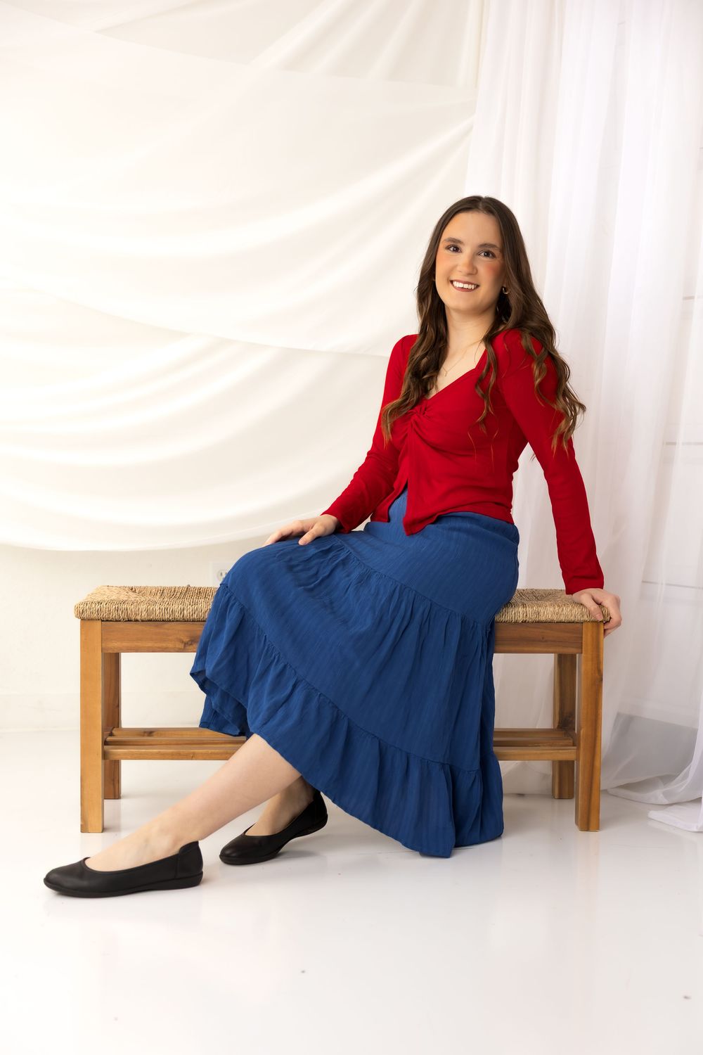 Woman in red top and blue midi skirt sits gracefully on a wooden bench against a white draped backdrop.