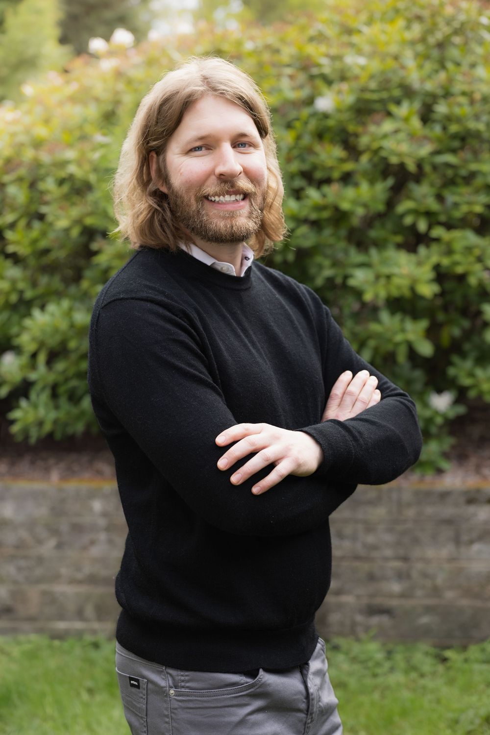 Smiling man with long blonde hair and beard, arms crossed, wearing black sweater outdoors in garden.