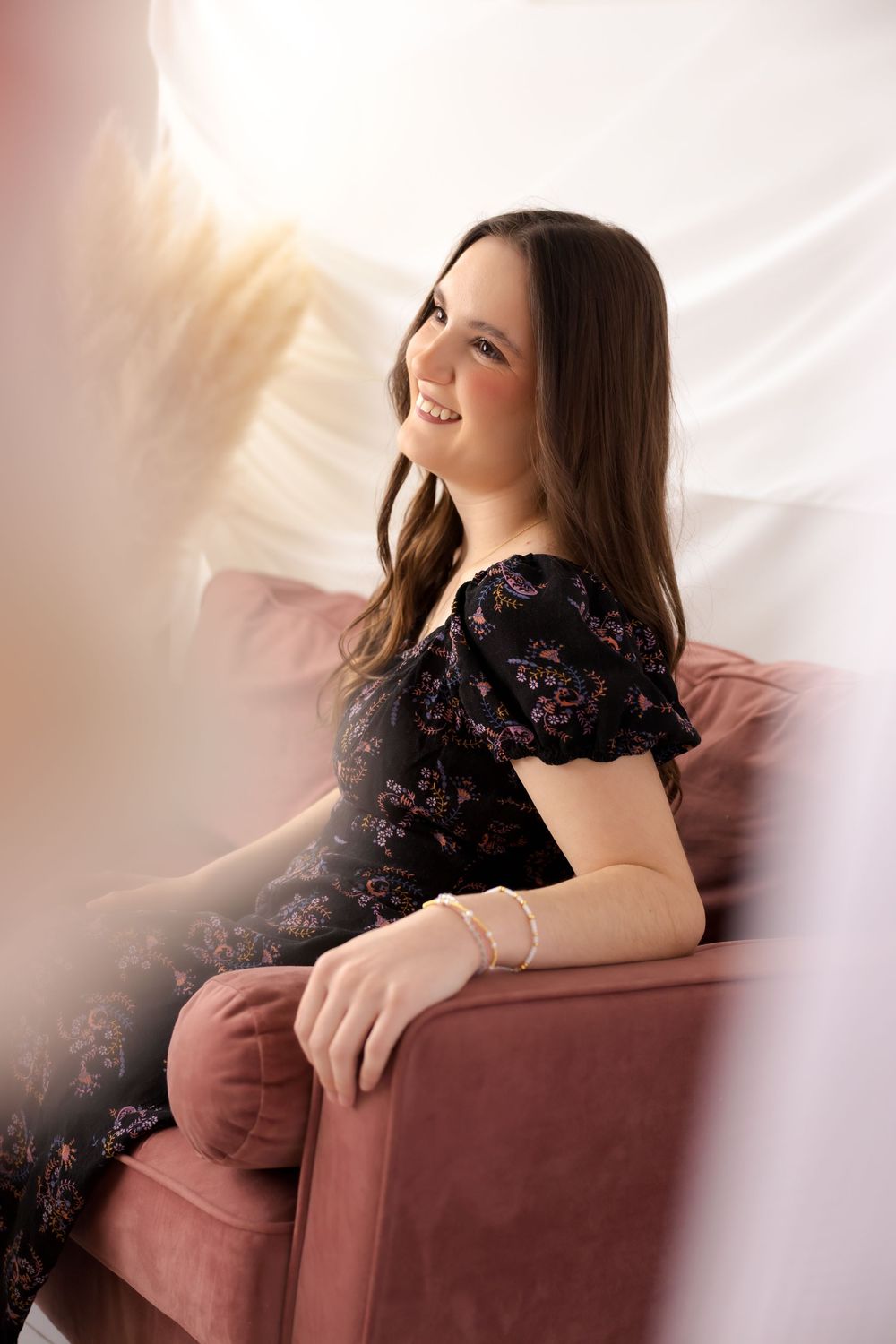 Young woman with long brown hair smiles while sitting on a pink velvet chair, wearing a black floral dress.