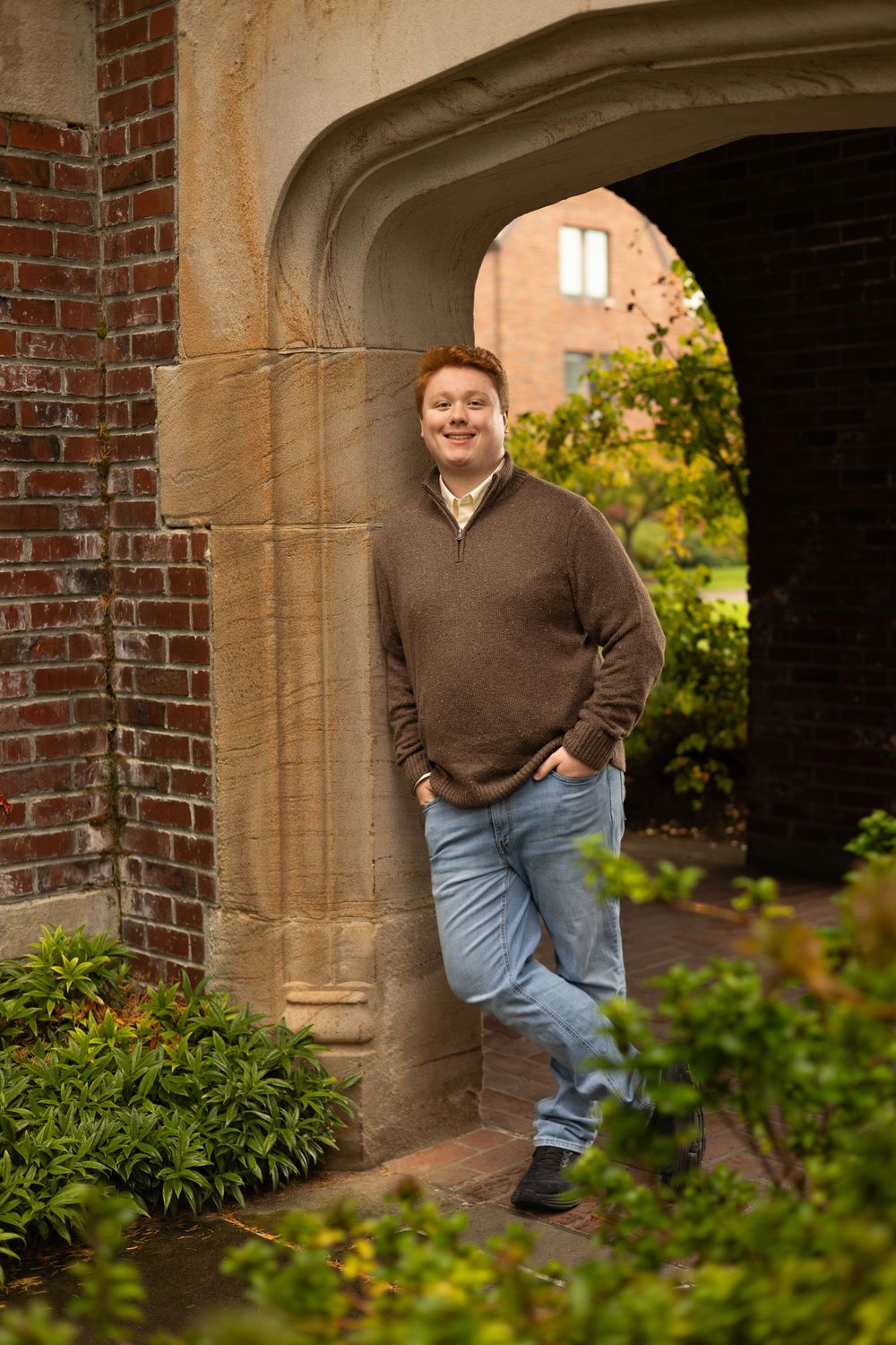 Person wearing brown sweater and jeans poses casually against brick archway surrounded by greenery.  Lifestyle Family Photography Tacoma WA