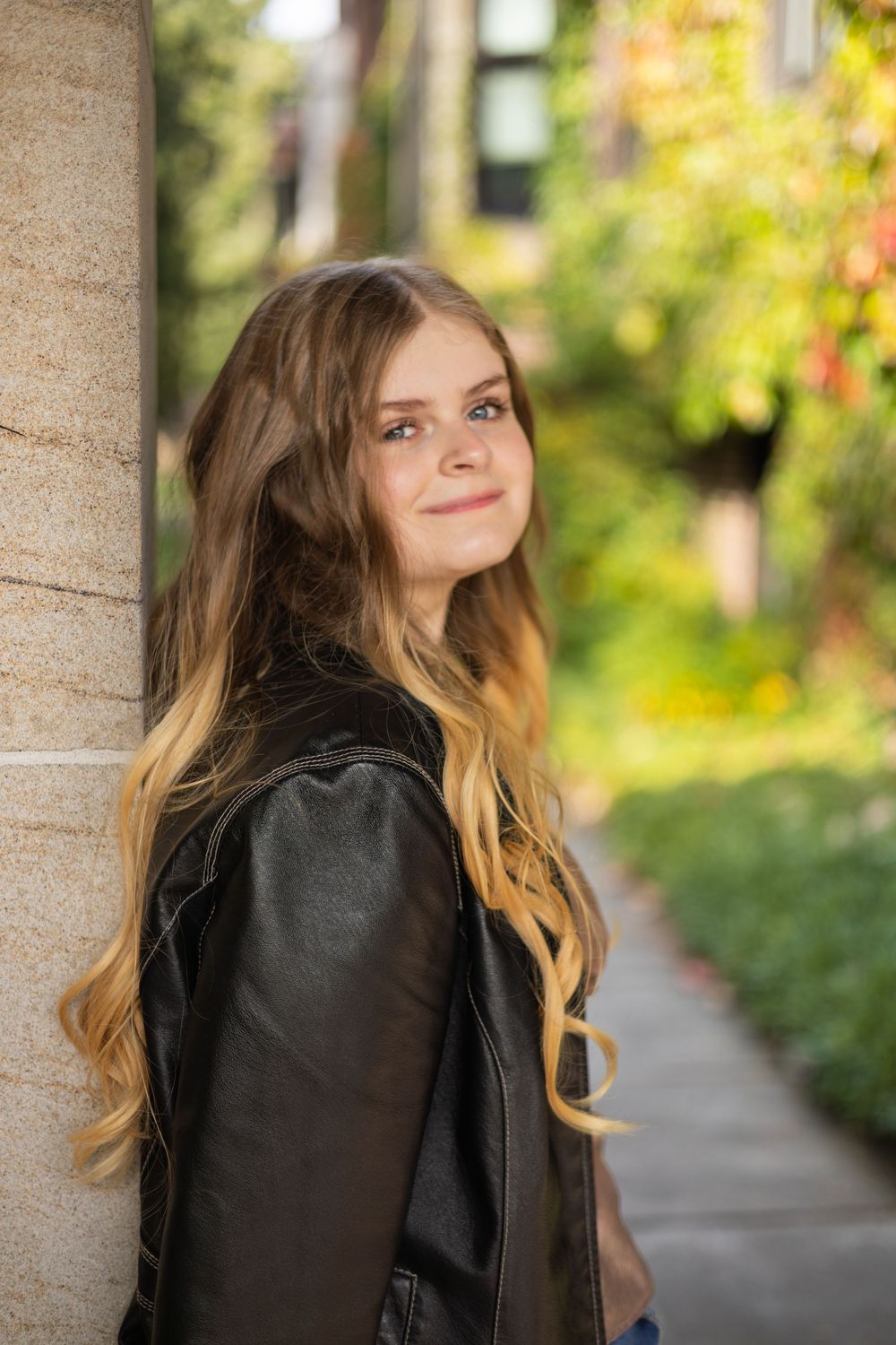 A person with long blonde hair poses in a black jacket against a sunlit garden background with autumn foliage.  Lifestyle Family Photography