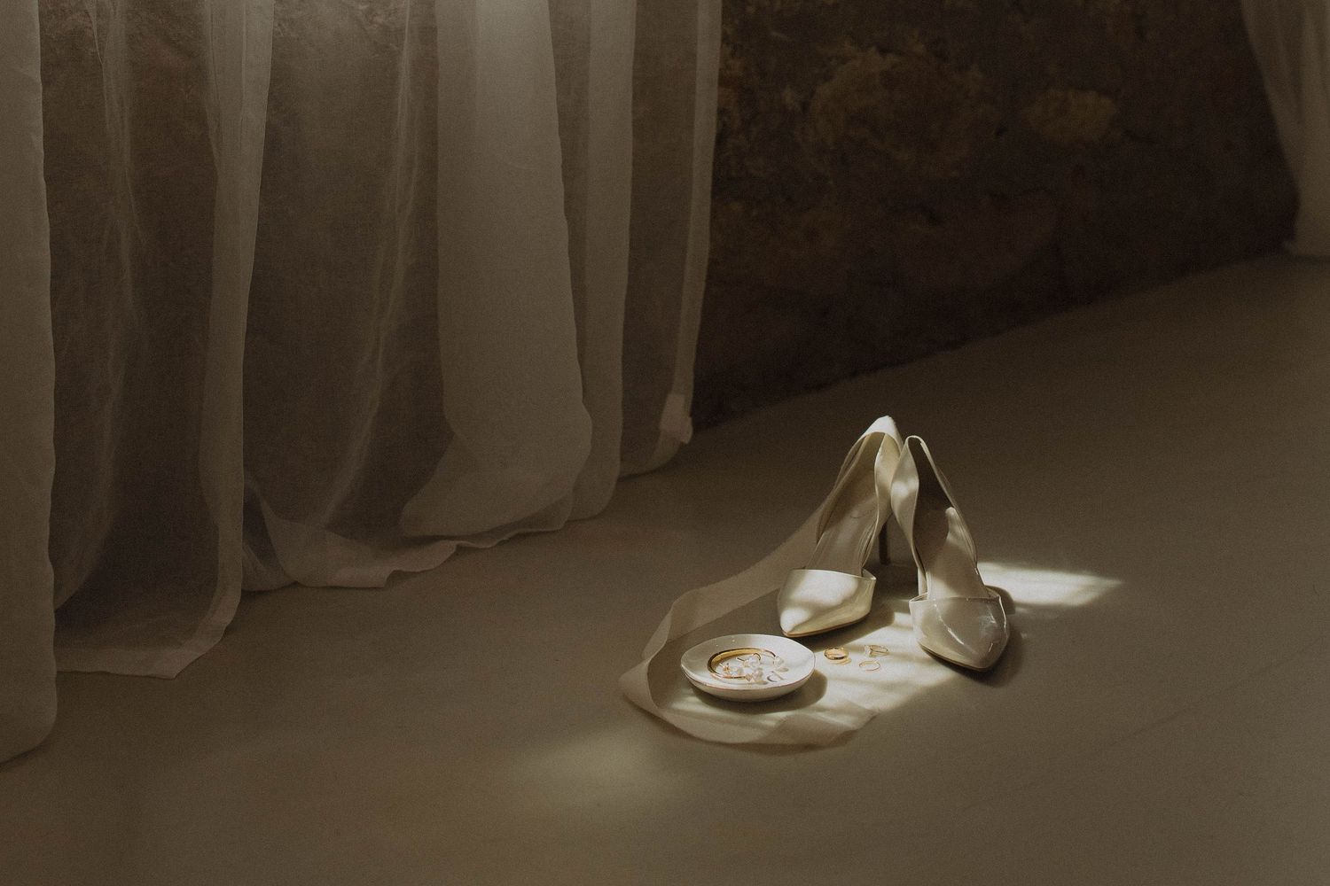 Wedding shoes arranged artistically on the floor against soft drapery in dim lighting.