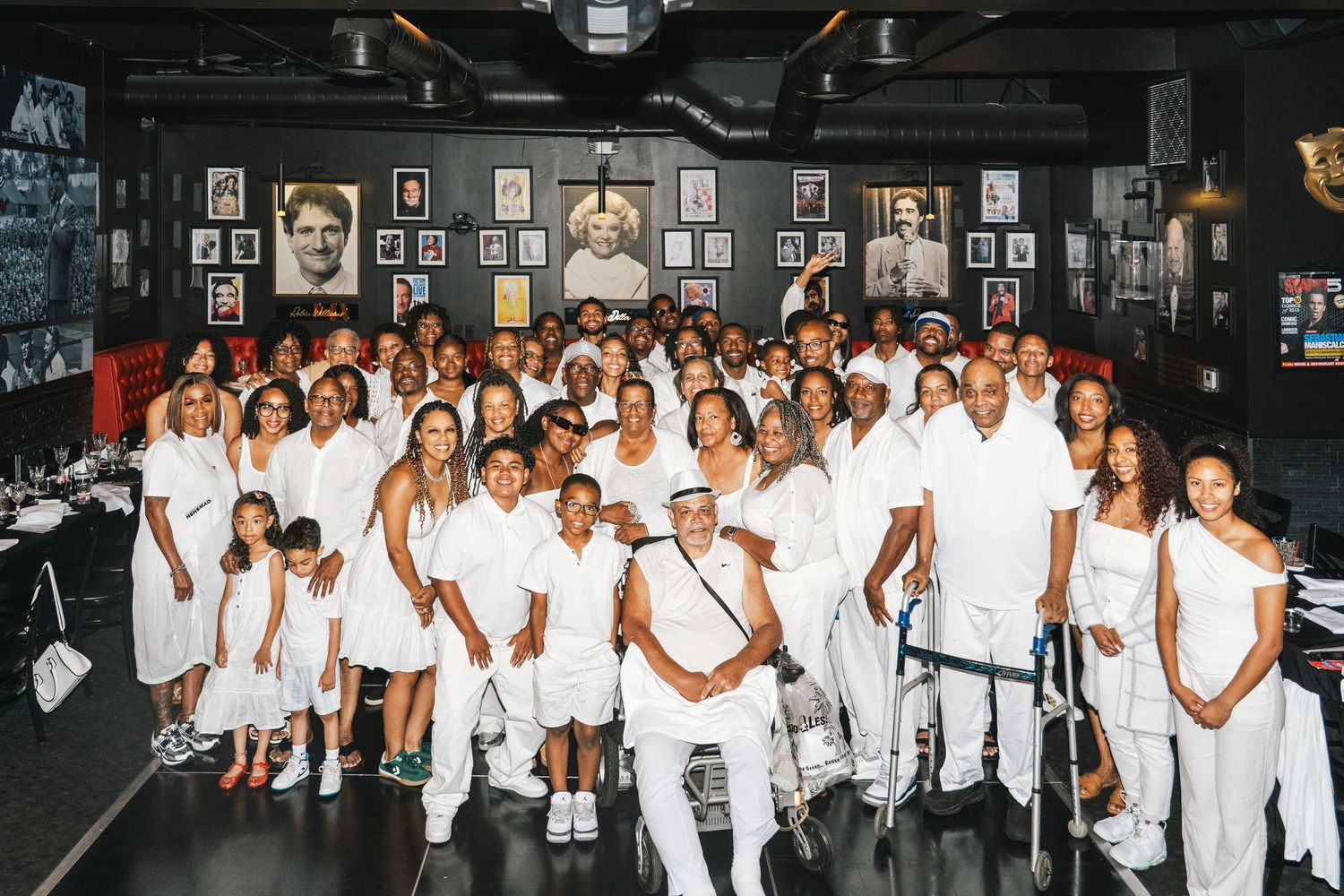 Large group photo of people wearing all white clothing in a dimly lit venue with photo-covered walls.