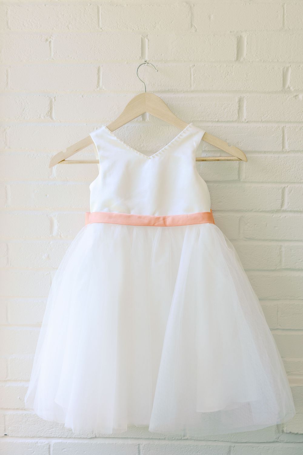 White flower girl dress with peach sash ribbon hanging against a light colored wall.