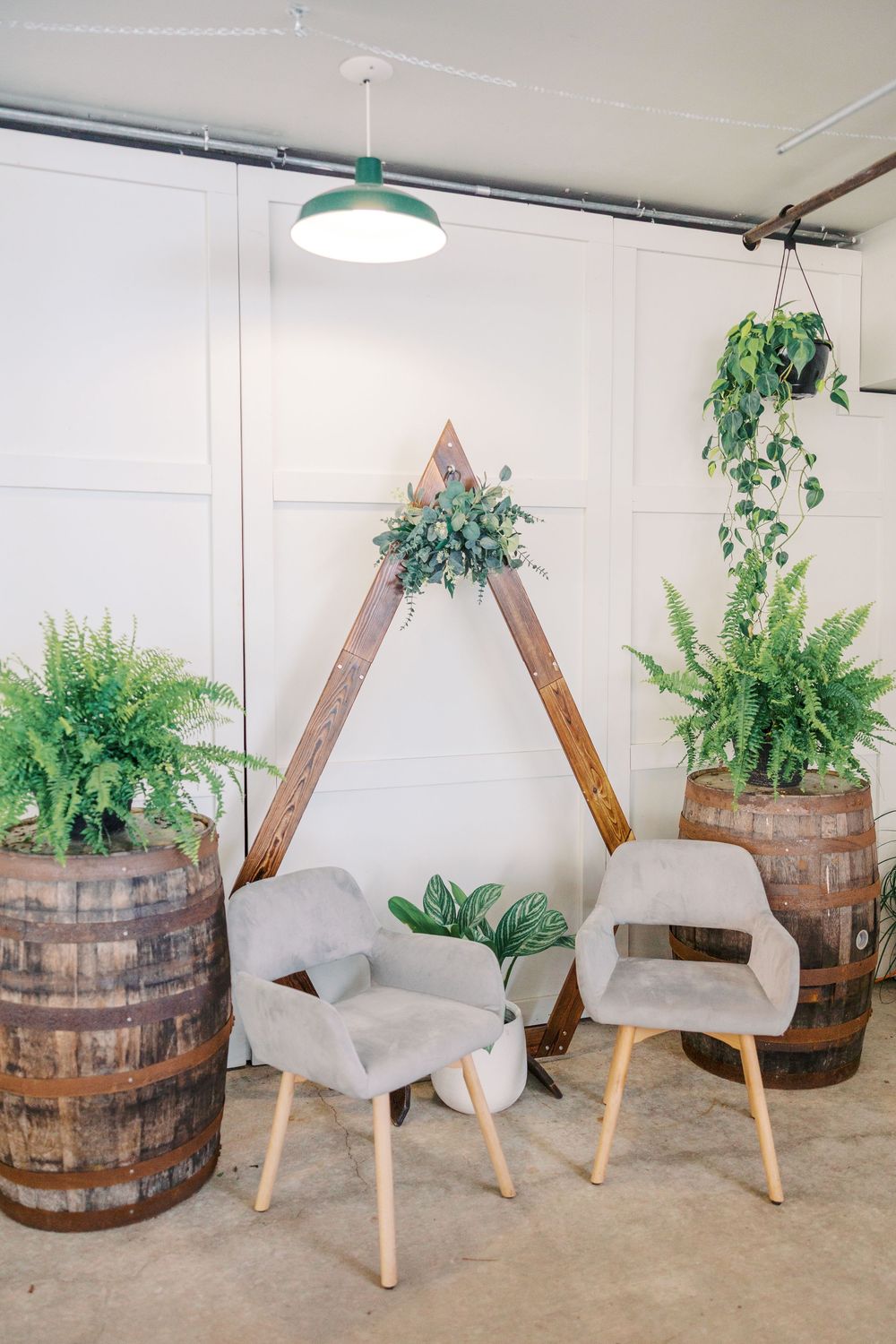Rustic wedding photo backdrop with wooden barrels, ferns and modern gray chairs arranged in front of a triangular frame.