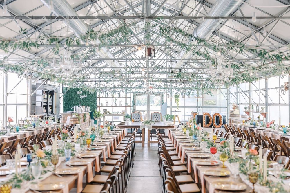 A bright indoor reception venue with long tables decorated with greenery and elegant place settings in a glass-enclosed space.