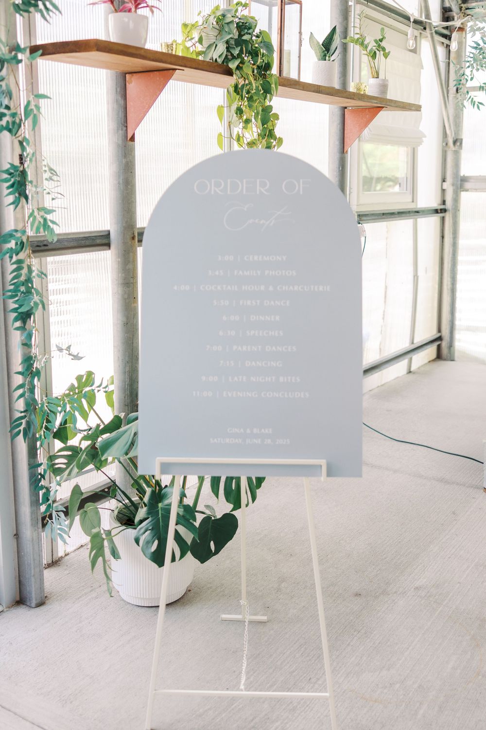 An elegant arched wedding order of events sign displayed on a white easel with greenery decor.