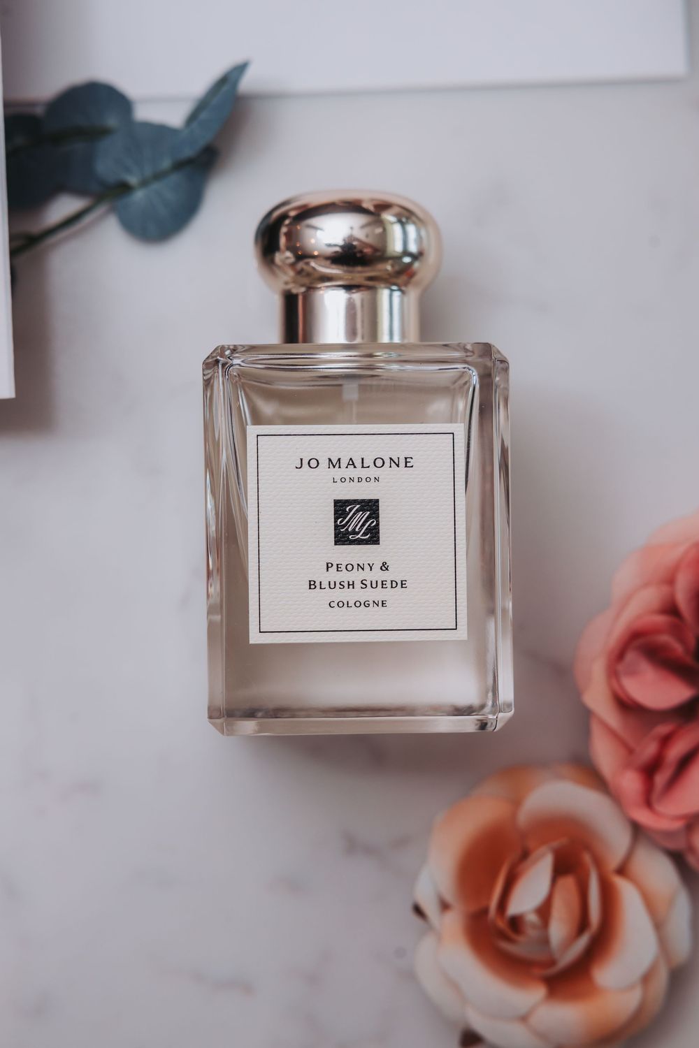 A Jo Malone Peony & Blush Suede cologne bottle staged with coral roses and eucalyptus on a marble surface.