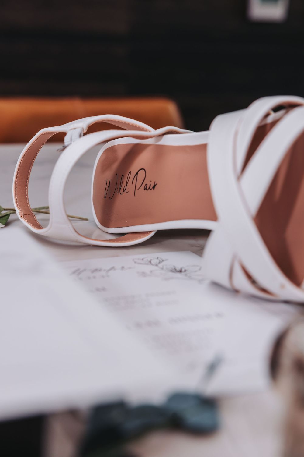 Close-up detail of white leather strappy sandals with signature branding on pink insole resting on paper surface.
