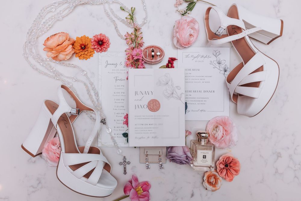 Wedding invitation suite with white heels and pink and coral flowers on a marble surface.