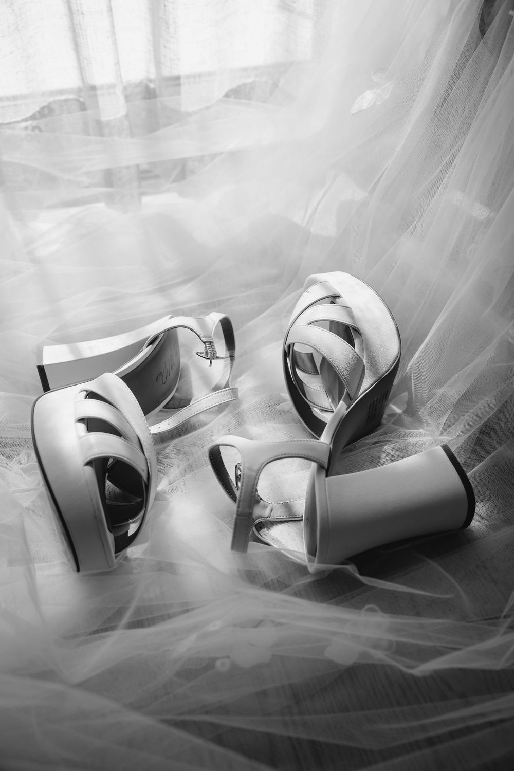 Black and white photo of elegant wedding heels resting on delicate tulle fabric in soft natural light.