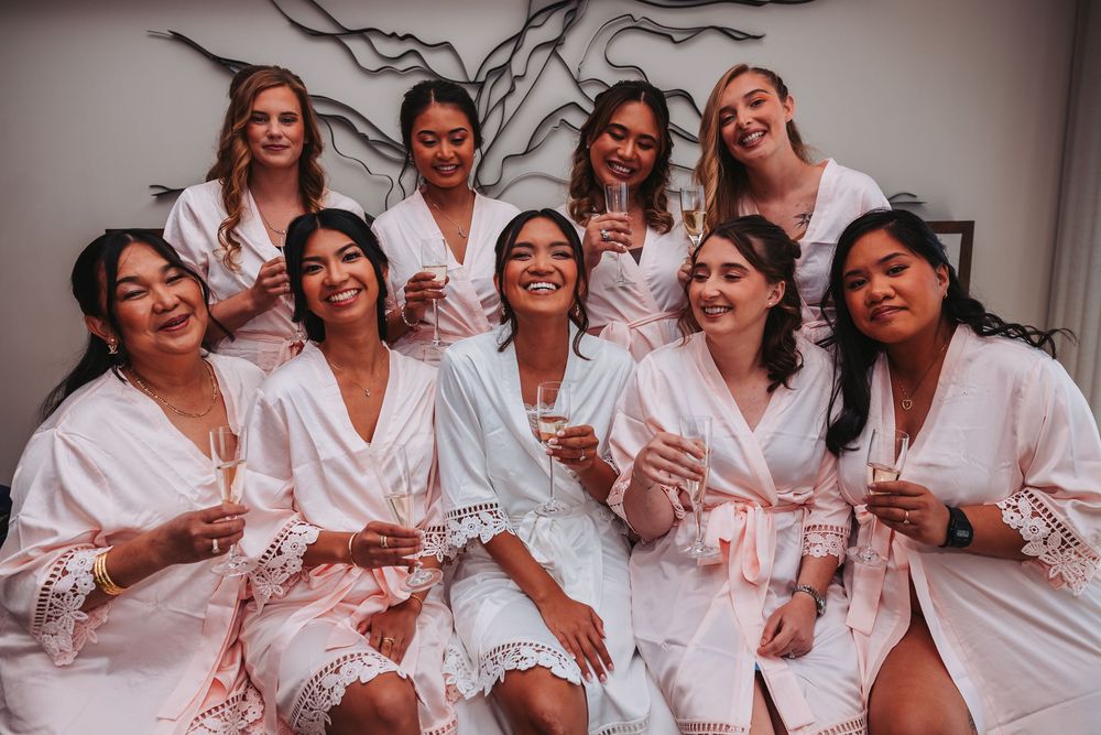 A group of women in matching pink satin robes laugh together during a wedding preparation photoshoot.