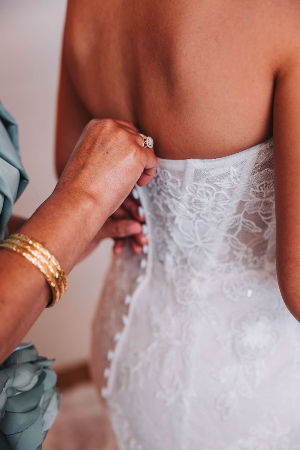 Close-up details of hands helping to fasten a delicate white lace wedding dress with ornate bracelets visible.