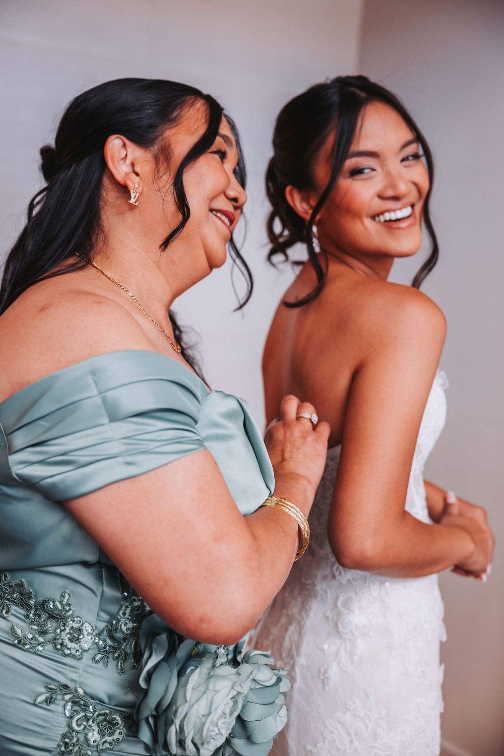 Two people share a joyful moment in formalwear, one in a sage green off-shoulder dress and one in a white bridal gown.