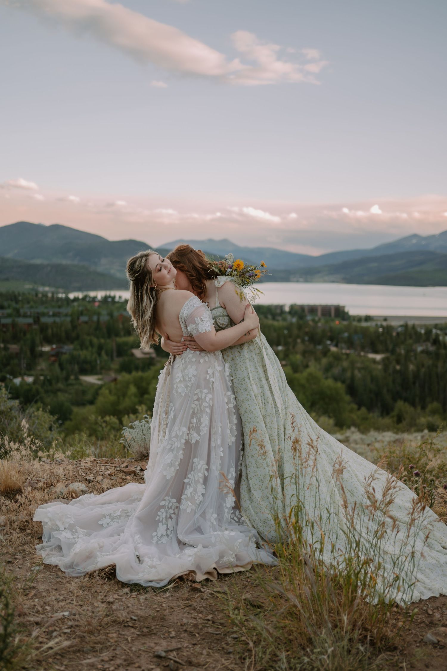 Two brides in wedding dresses embrace on a hilltop with a scenic view of mountains, a lake, and a town below.
