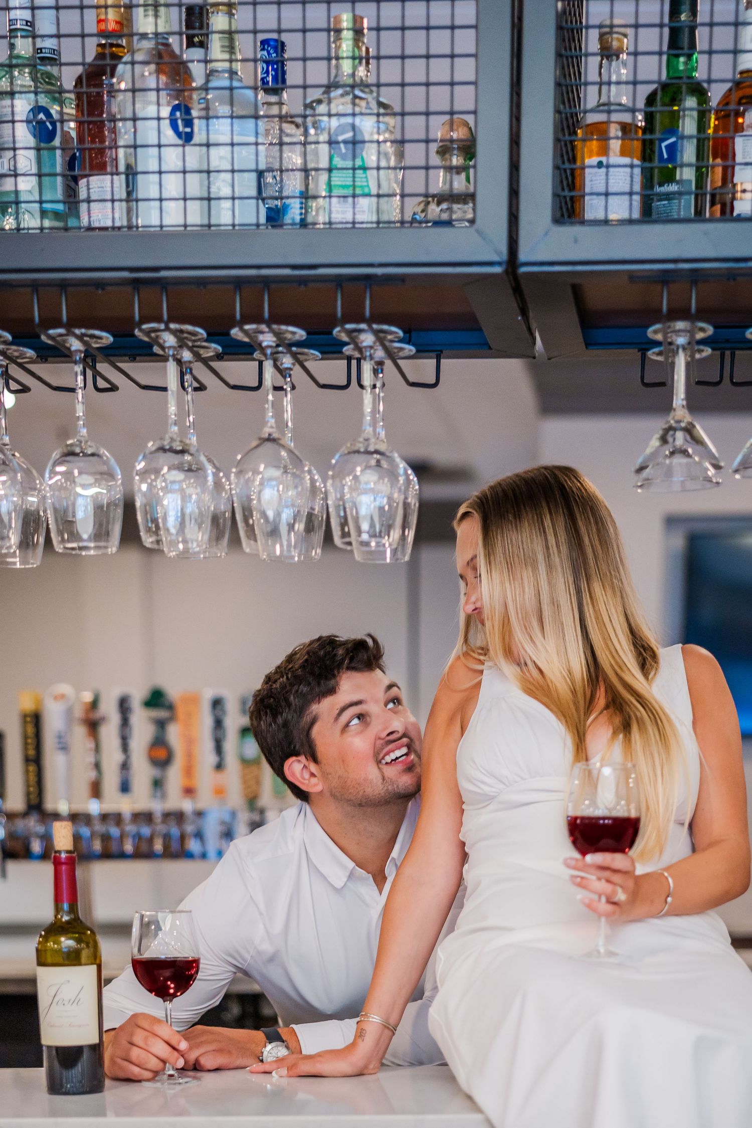A couple enjoys wine at a stylish bar with hanging wine glasses and bottles displayed on shelves above.