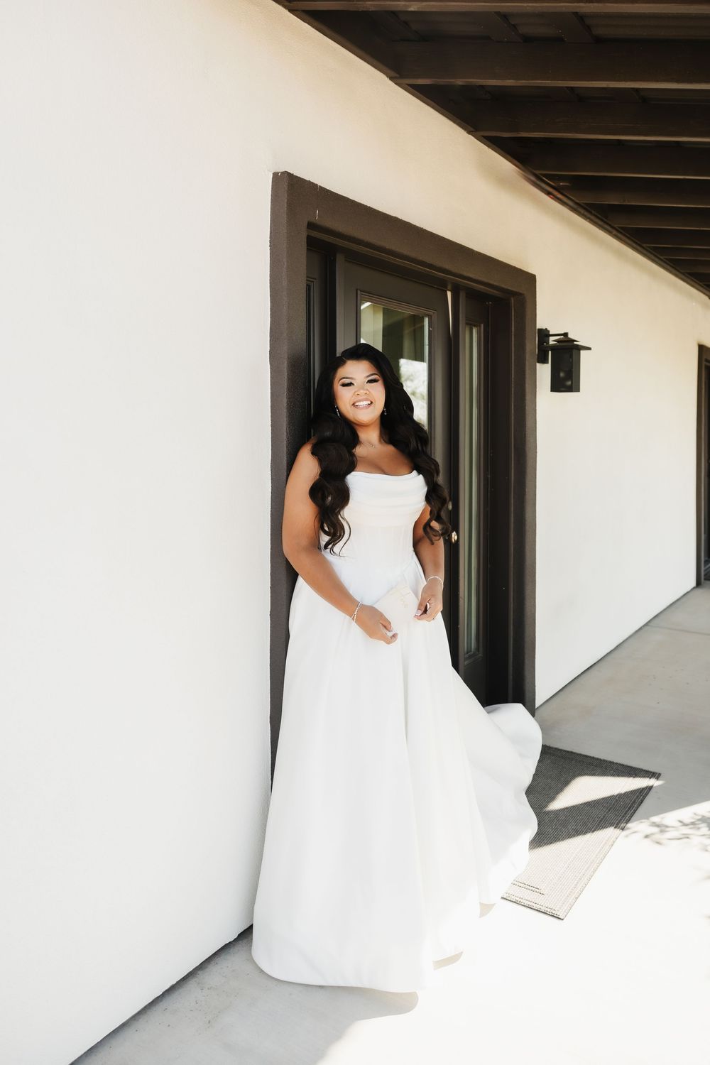 A bride in a white wedding dress poses on a sunlit porch with modern architecture.