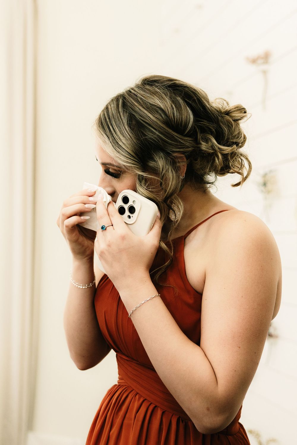 Person in burgundy dress has emotional reaction while holding phone during wedding preparations.