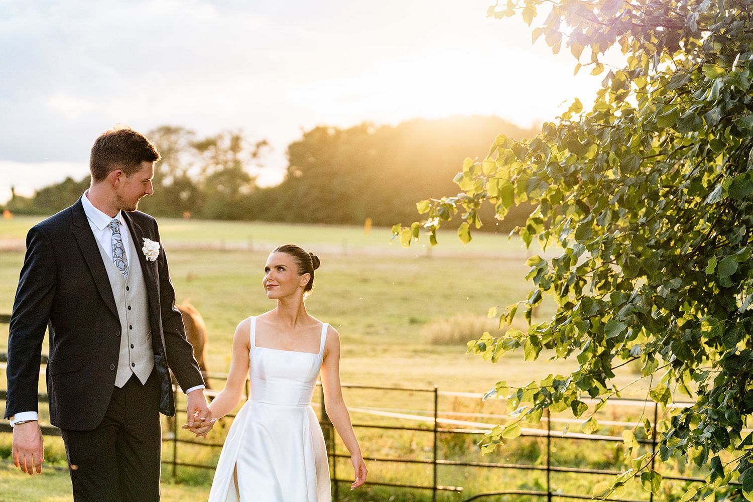 Newlyweds walk hand in hand through a sunlit countryside field at golden hour with rustic fence in background.