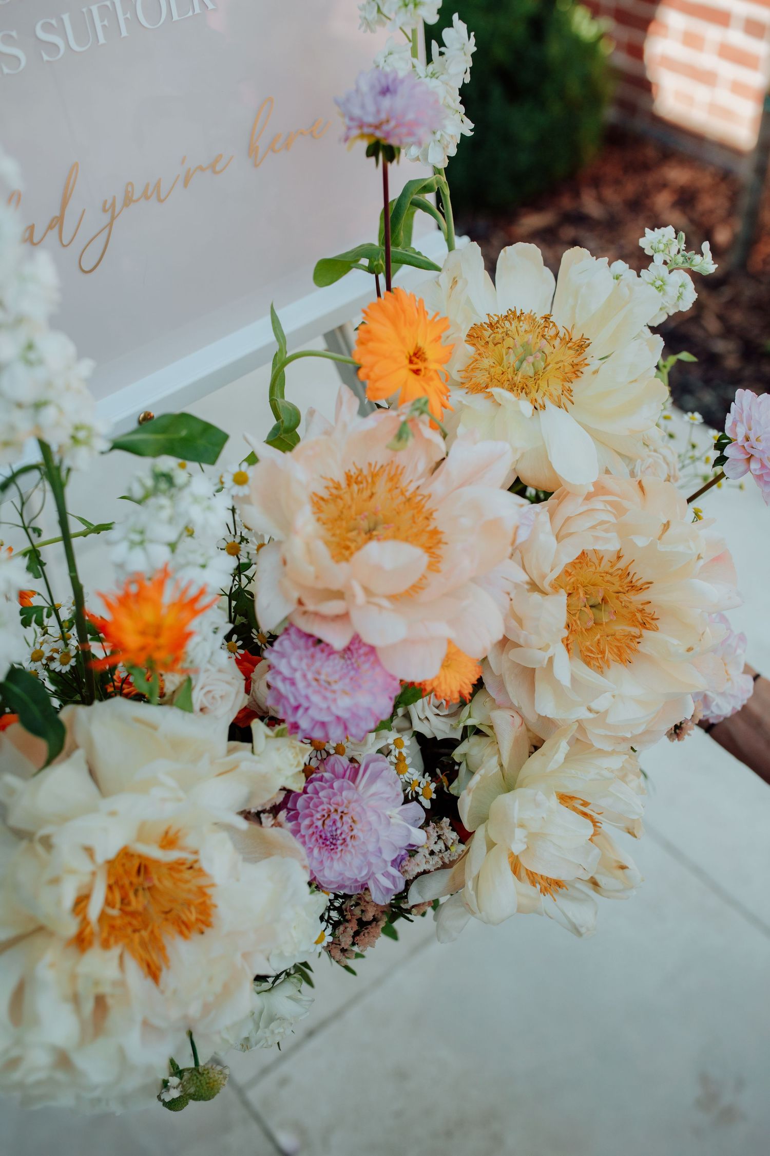 Close-up view of romantic floral arrangement with white peonies, orange marigolds, and pink accents on light marble surface.