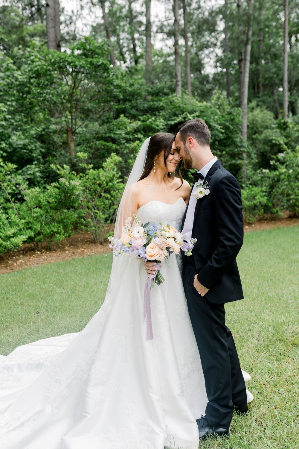 A couple shares an intimate moment in their wedding attire among trees in a lush garden setting.