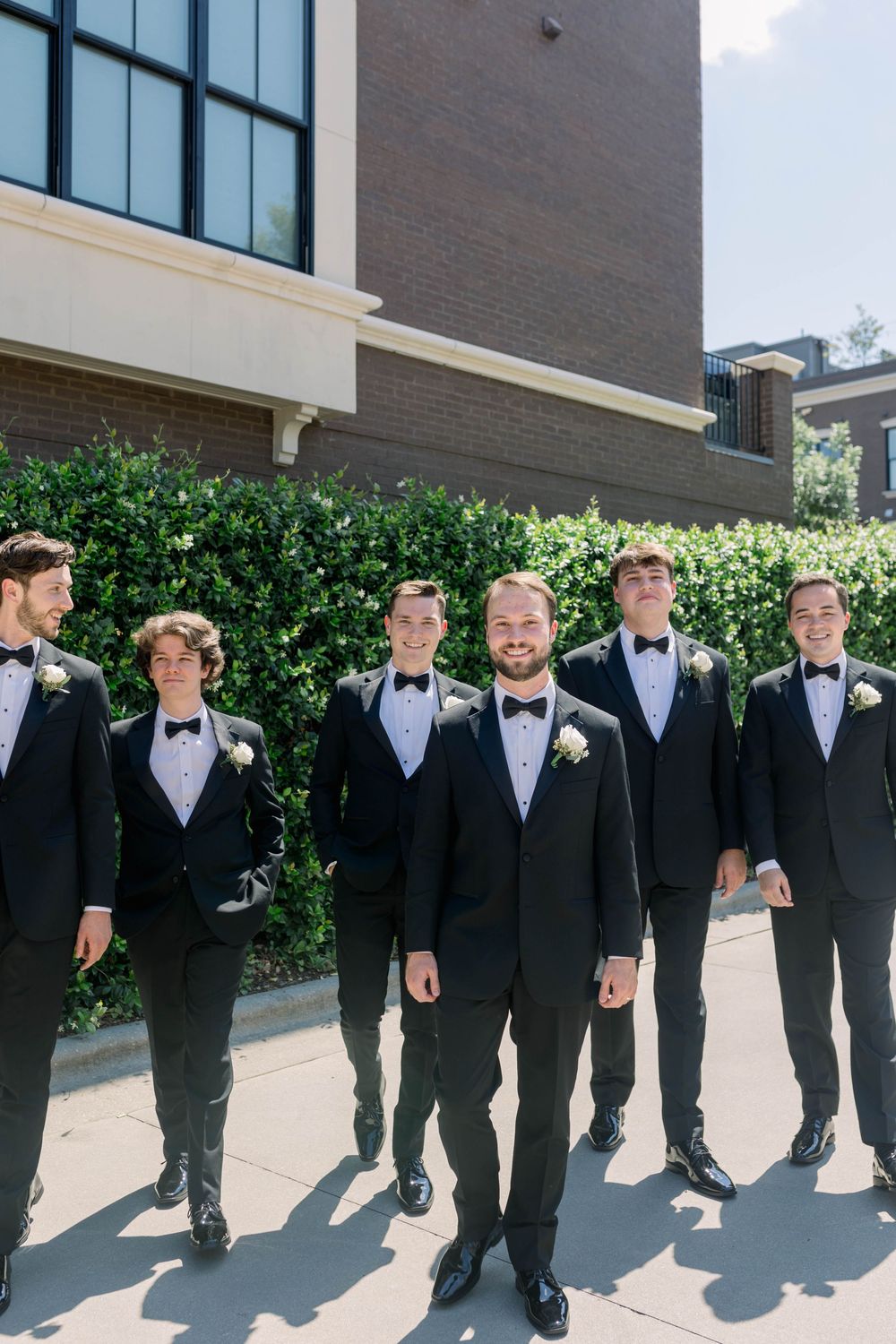 Groomsmen in black tuxedos and bowties line up against a building wall for a formal wedding photo.