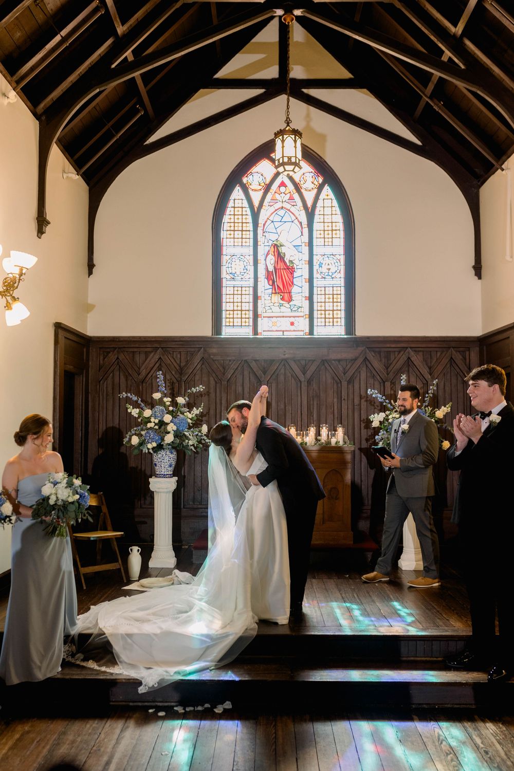 A wedding ceremony takes place in a historic chapel with exposed wooden beams and stained glass windows.