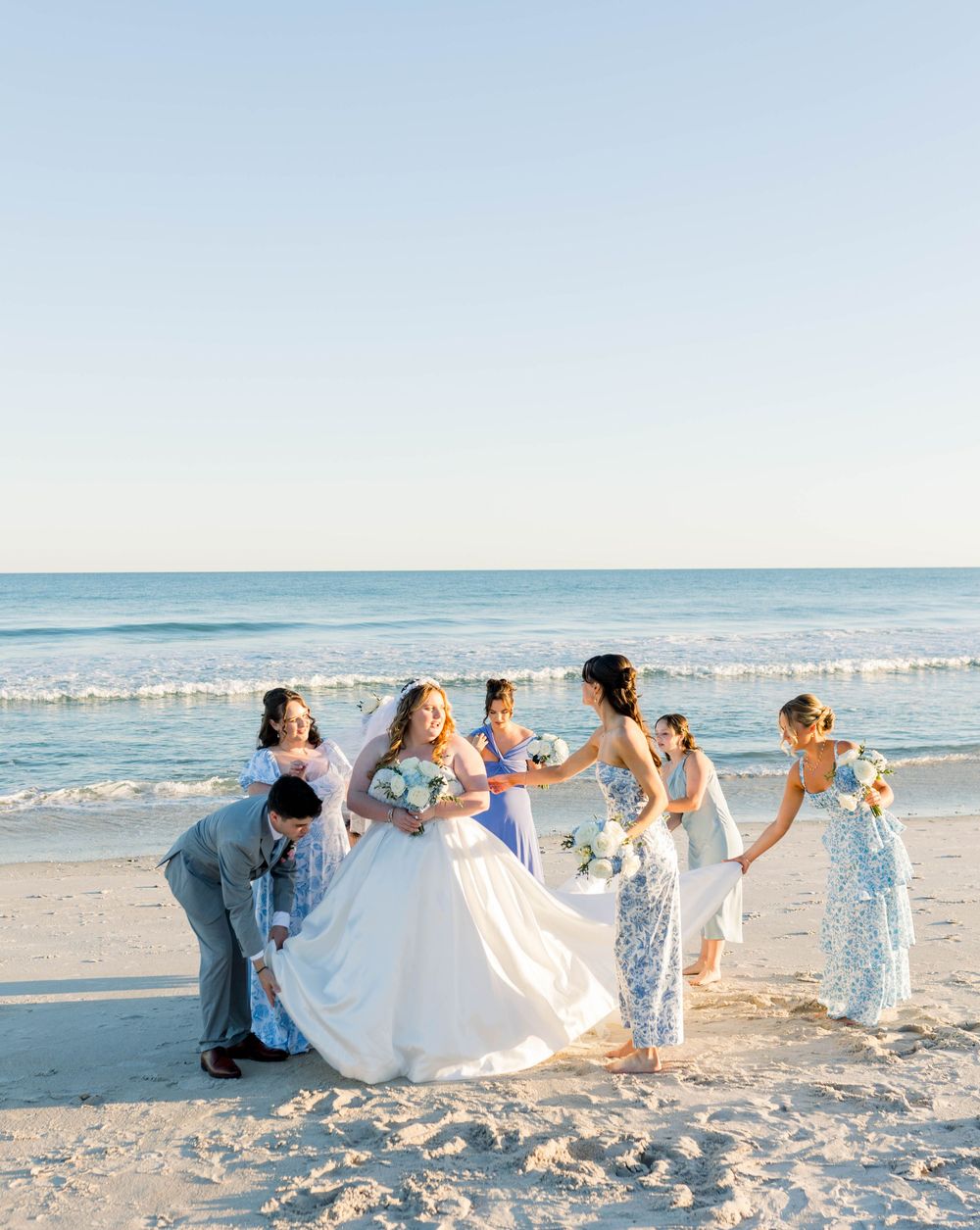 Wedding party celebrates on a sunny beach with the bride's white dress flowing in the ocean breeze.