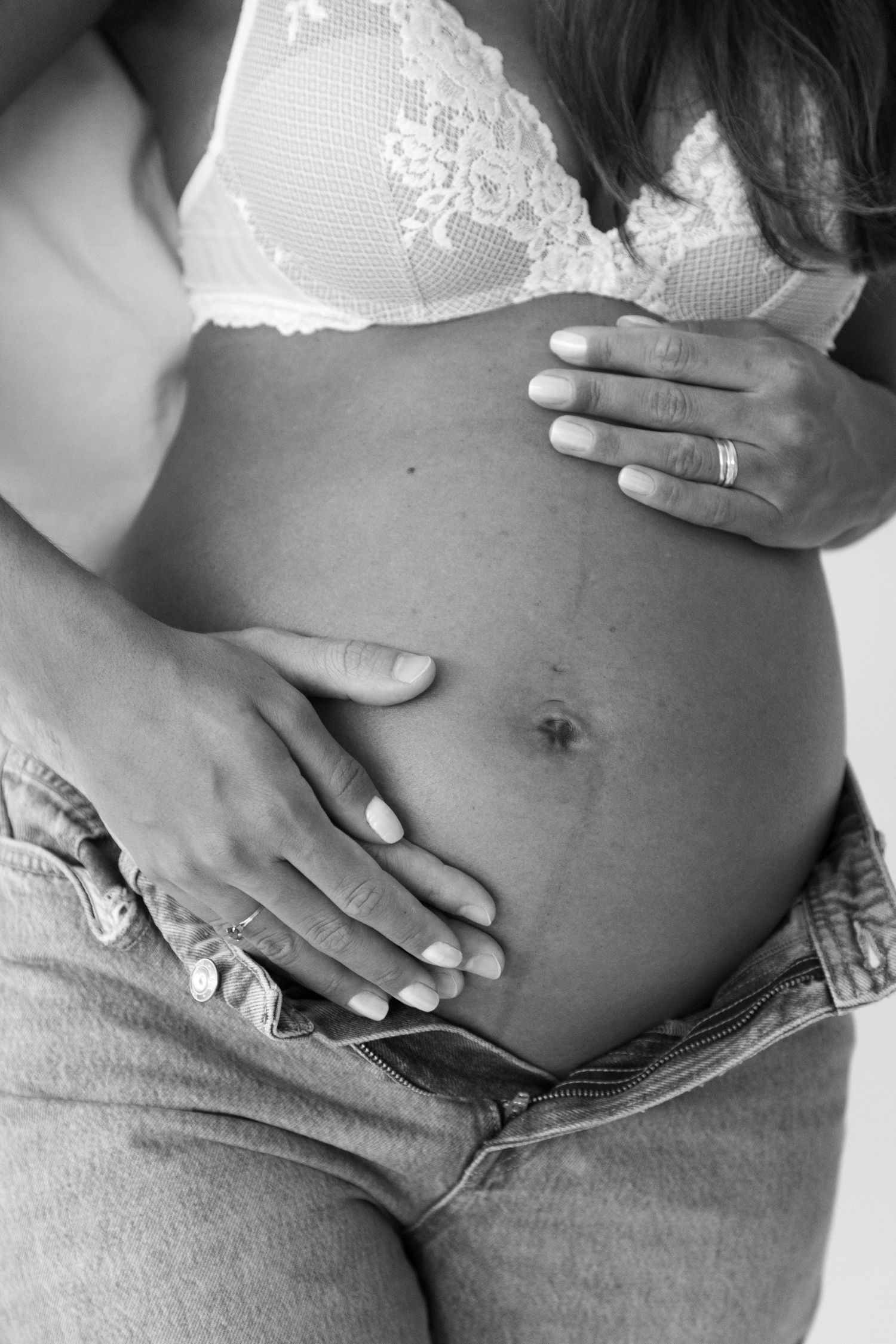 Black and white photo of a pregnant belly with hands gently resting on it, wearing white lace bra and jeans.