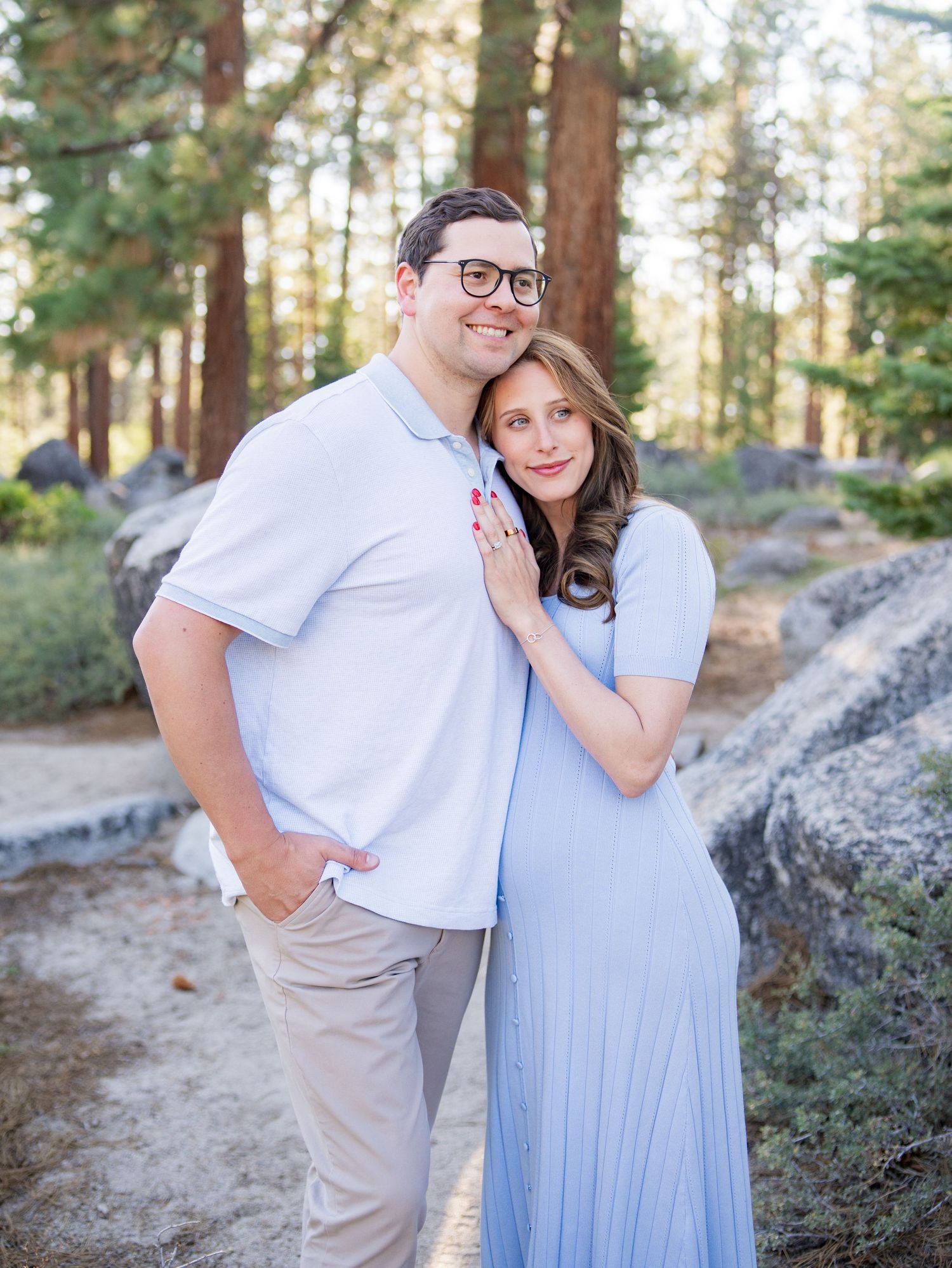 A romantic maternity photo showing a couple sharing a kiss against granite boulders in a forest setting.