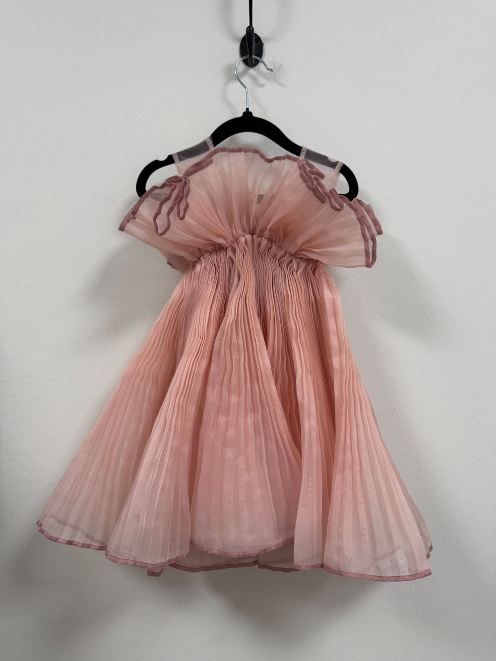 A delicate blush pink pleated chiffon dress with flutter sleeves hangs elegantly on a black hanger against a white wall.