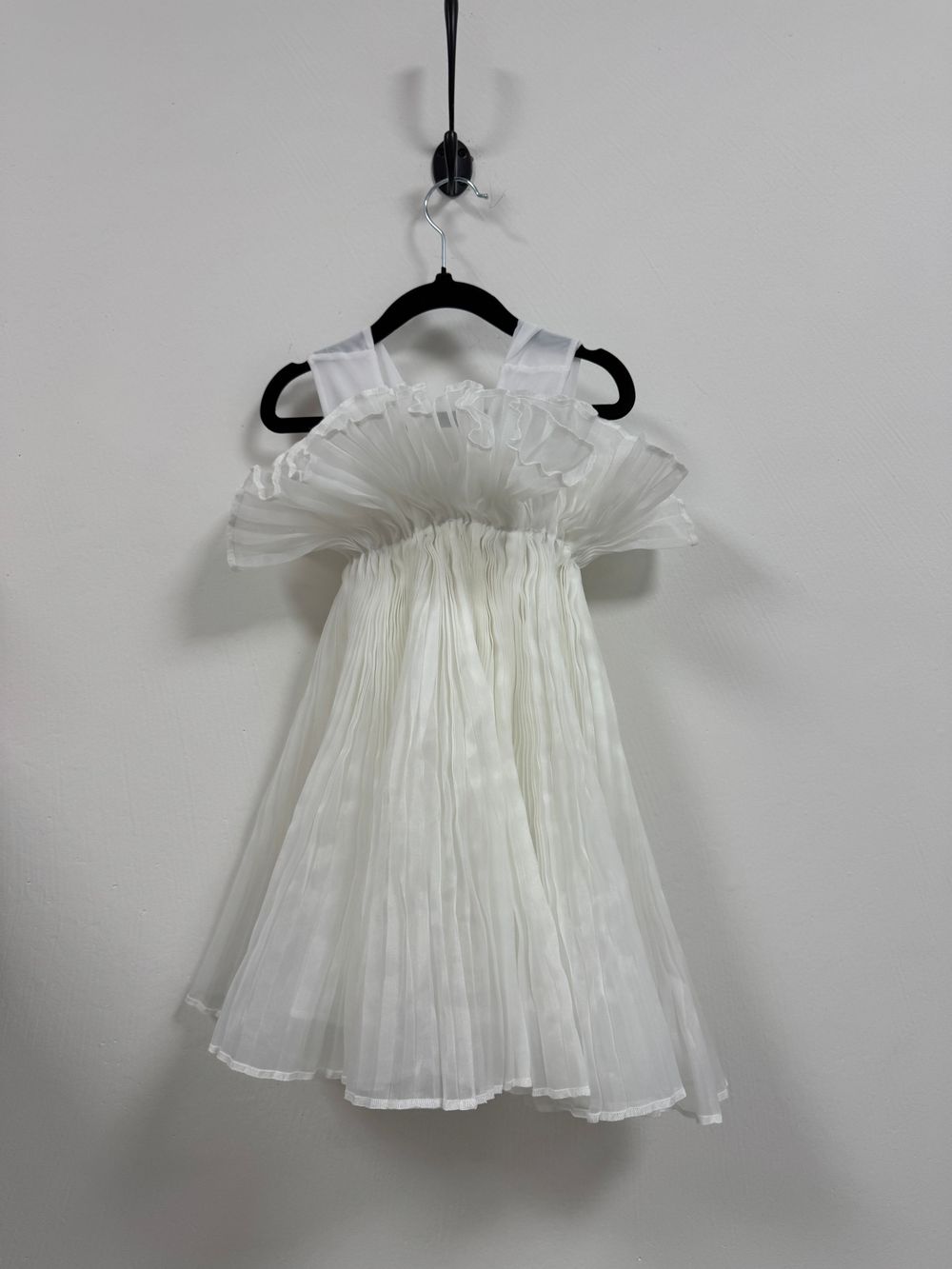 A delicate white pleated sleeveless dress with ruffled shoulders hangs on a black hanger against a plain wall.