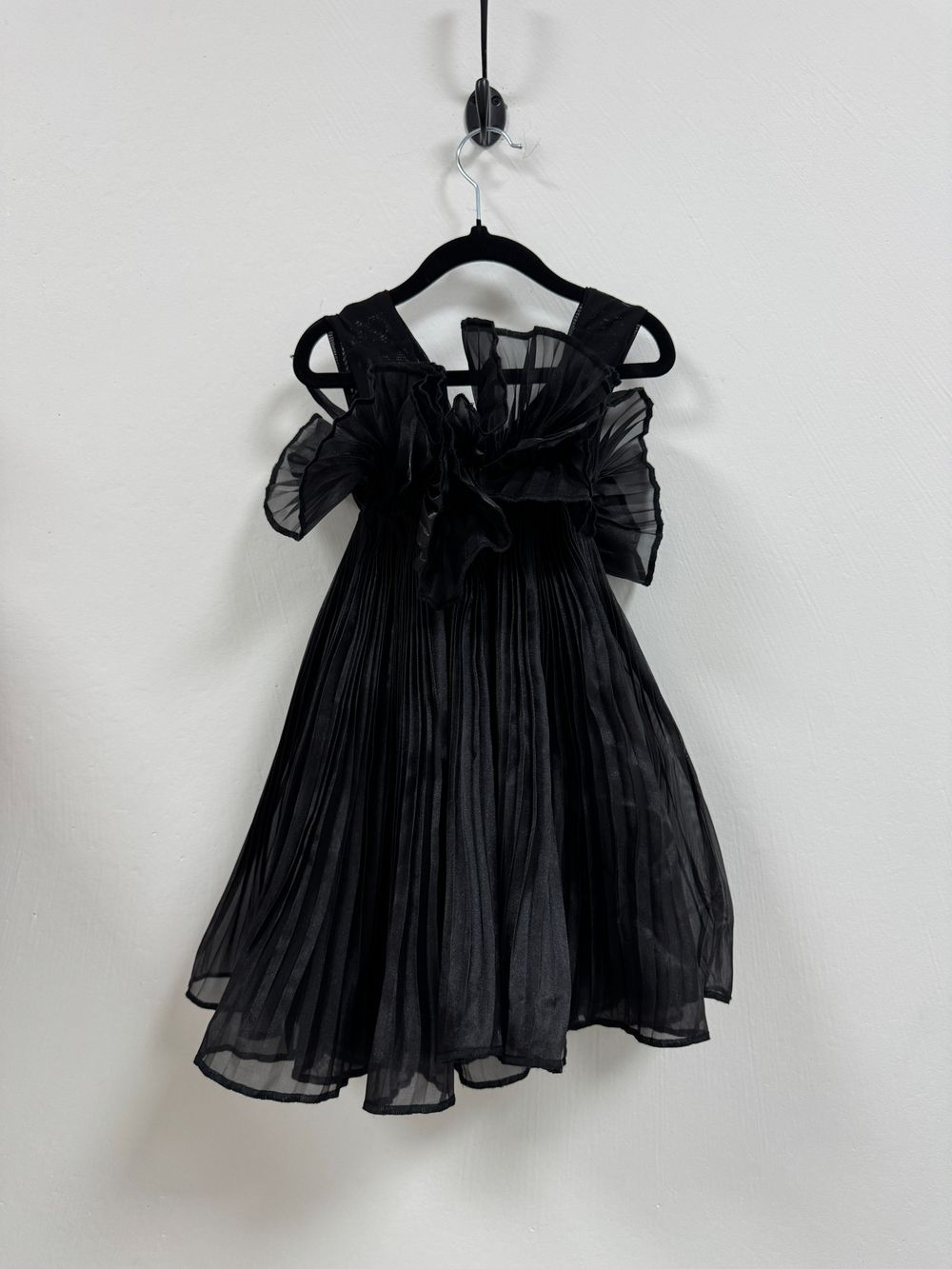 A black tulle party dress with gathered skirt and bow detail hangs on a white wall from a wooden hanger.