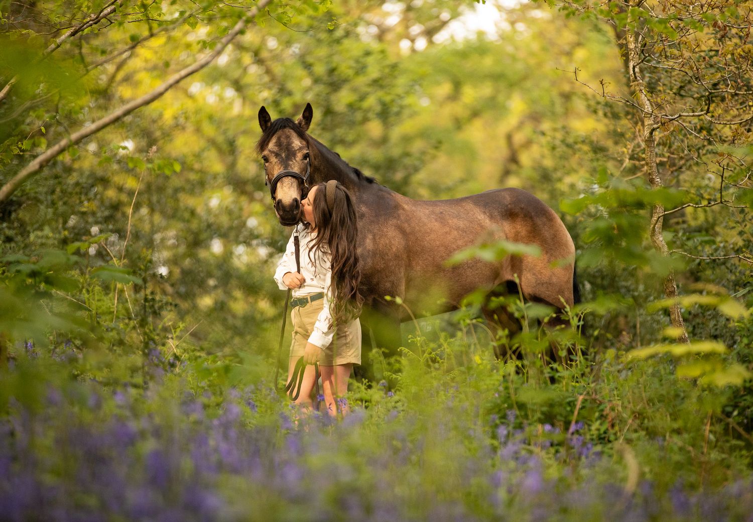 A brown horse and someone stand together in a forest clearing with purple wildflowers blooming around them.