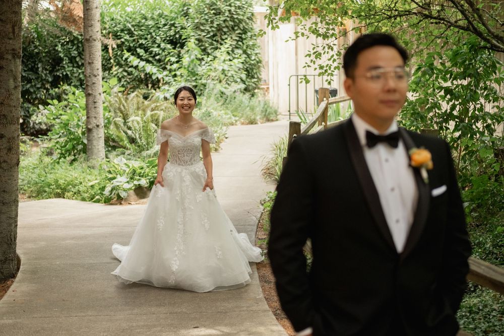 Bride in white ball gown approaches groom in black tuxedo during outdoor wedding first look ceremony.