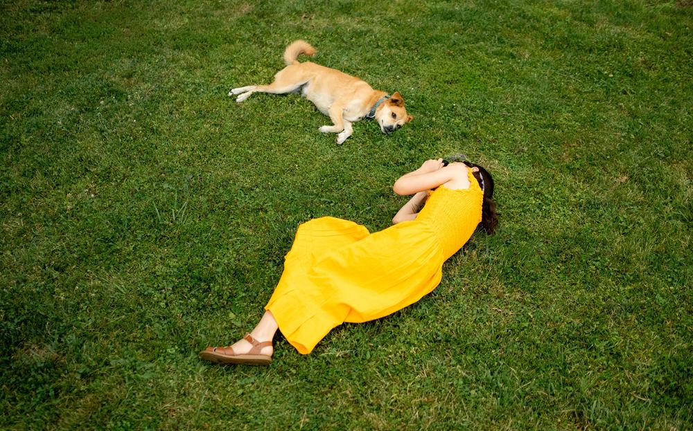 Person in yellow dress and dog laying on grass during sunny day.