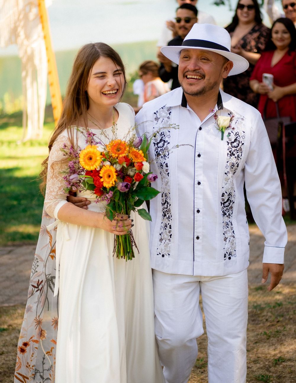 Wedding party in white attire holds vibrant orange and yellow bouquets outdoors.