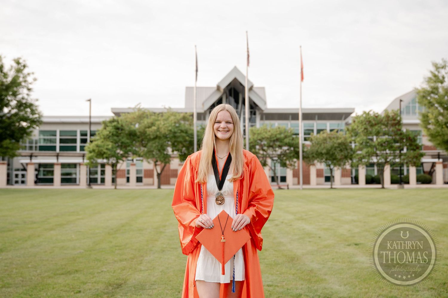 Graduate in bright orange academic regalia walks and poses for photos on a green lawn near campus buildings.