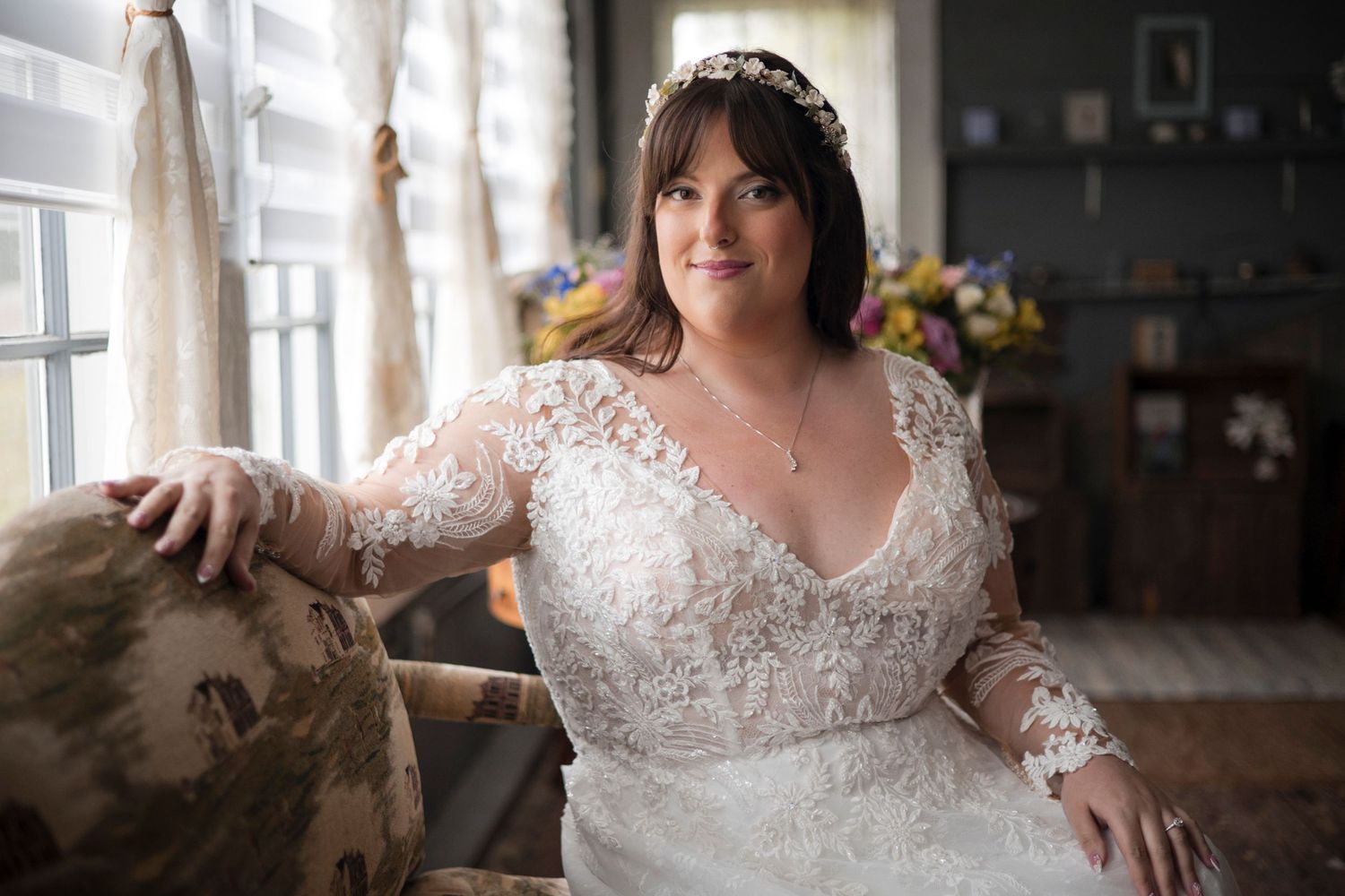 A bride in a lace long sleeve wedding gown poses near a window in a vintage-style interior setting.