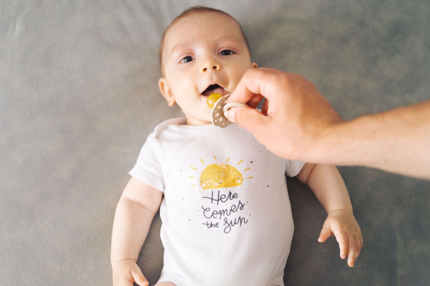 Baby wearing white onesie being fed with a spoon while lying on grey background.