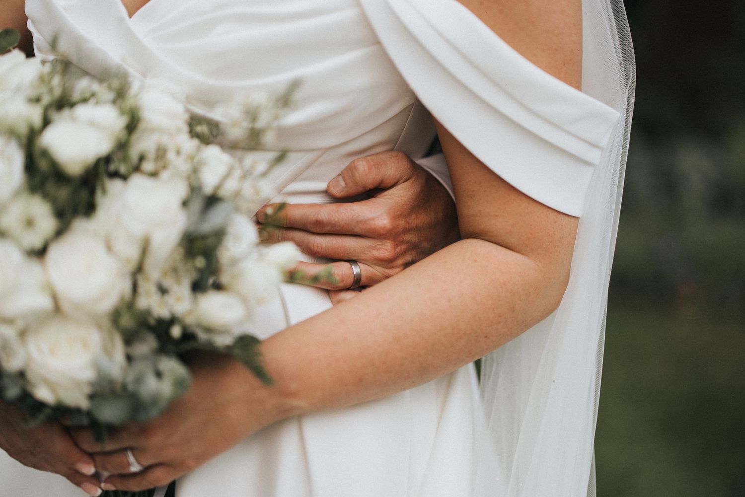 Close up detail of a wedding dress and white bouquet showing hands holding flowers and fabric draping elegantly.