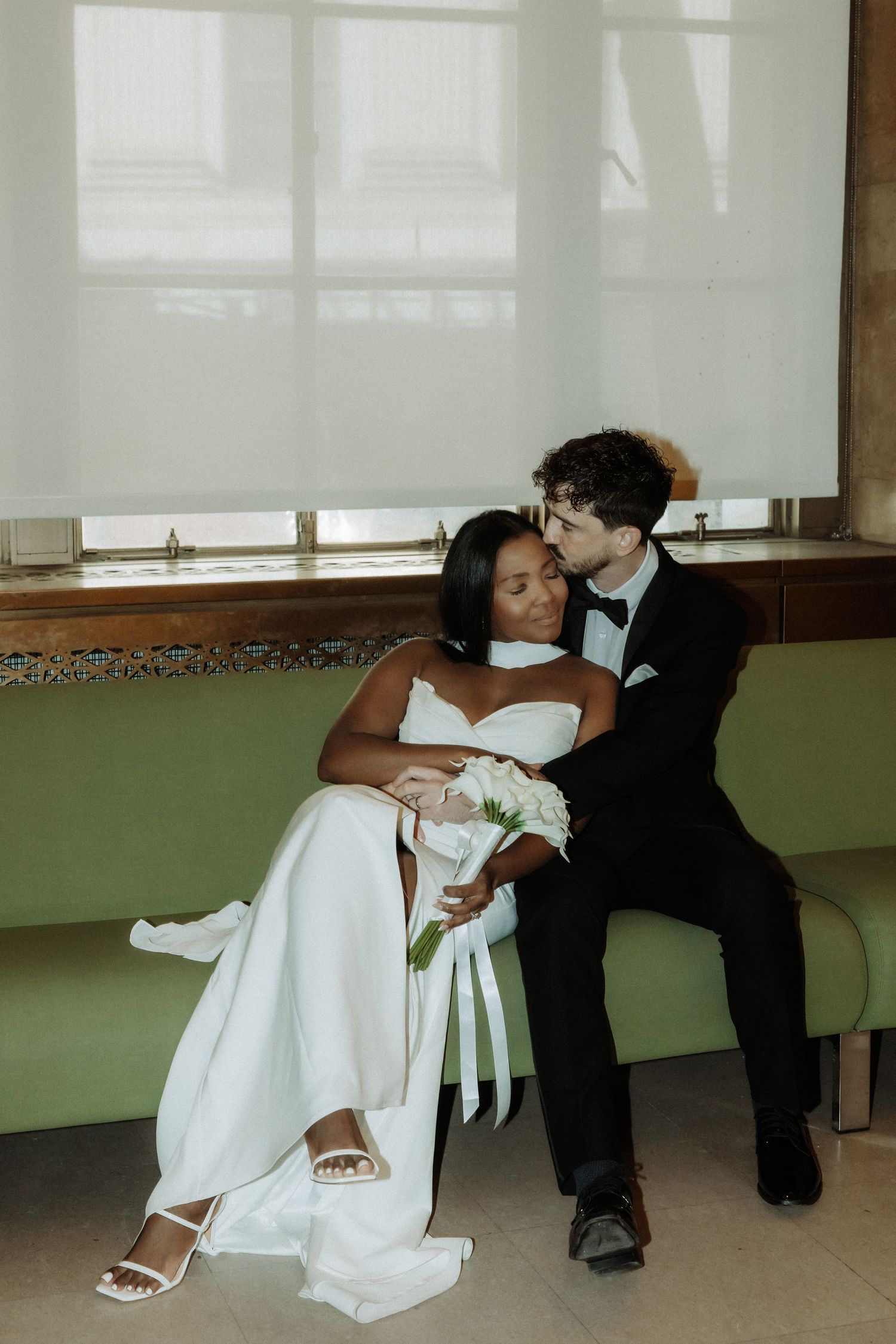 Newlyweds share a playful moment while sitting on a bench in an intimate indoor setting.