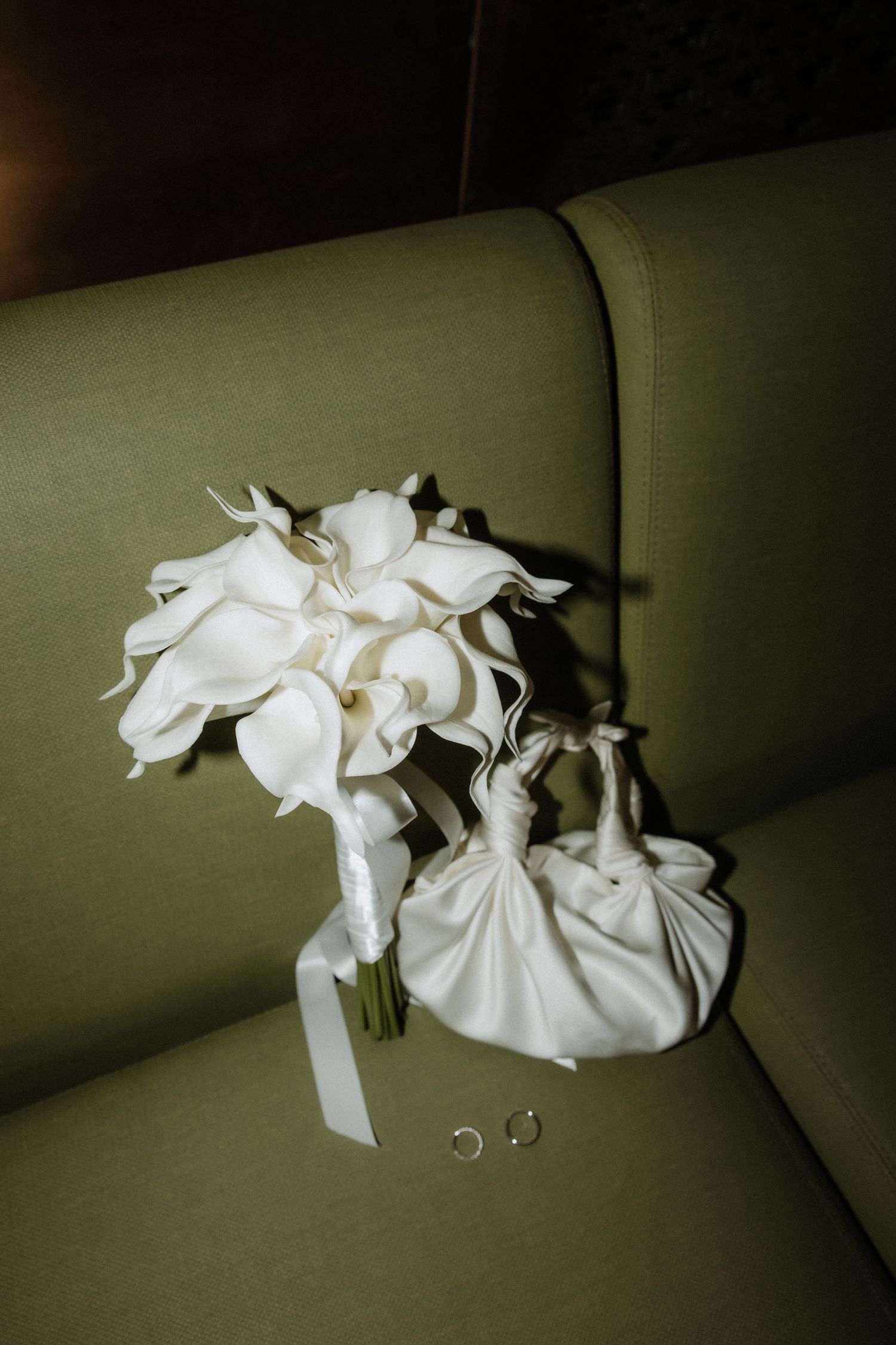 A white calla lily bridal bouquet and small purse resting on an olive green sofa.