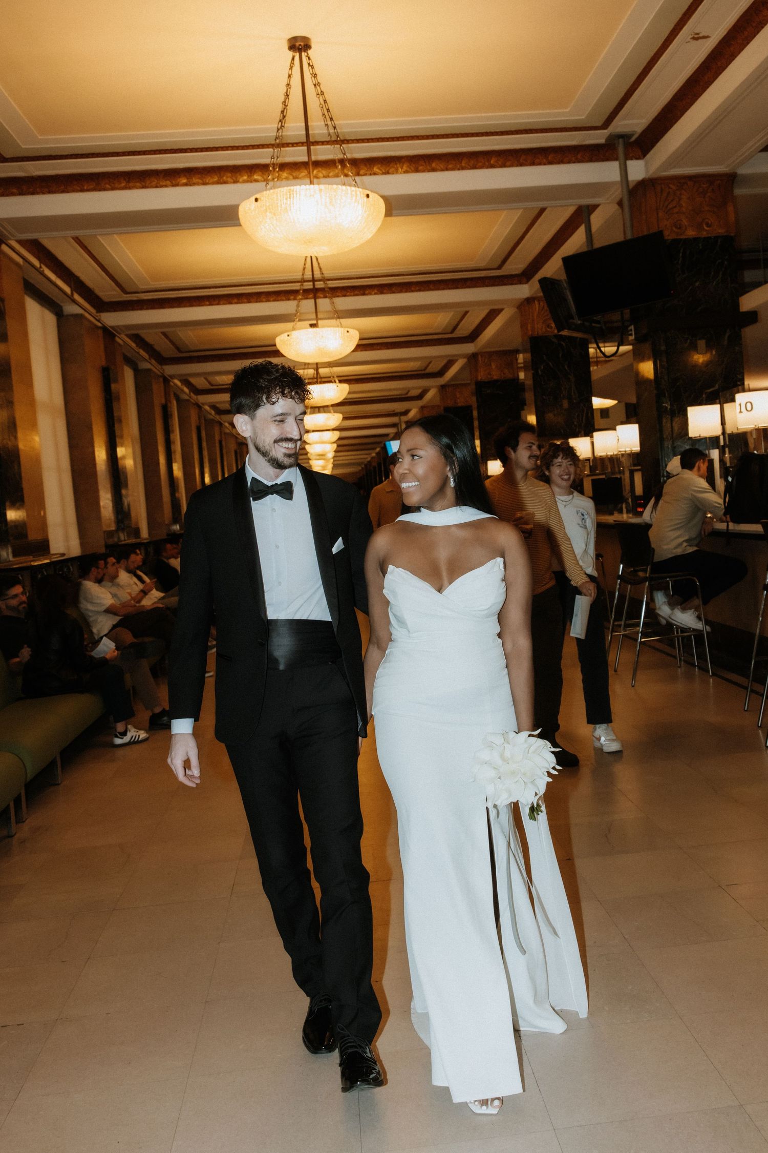 A couple in formal wedding attire dances across an elegant ballroom with crystal chandeliers.