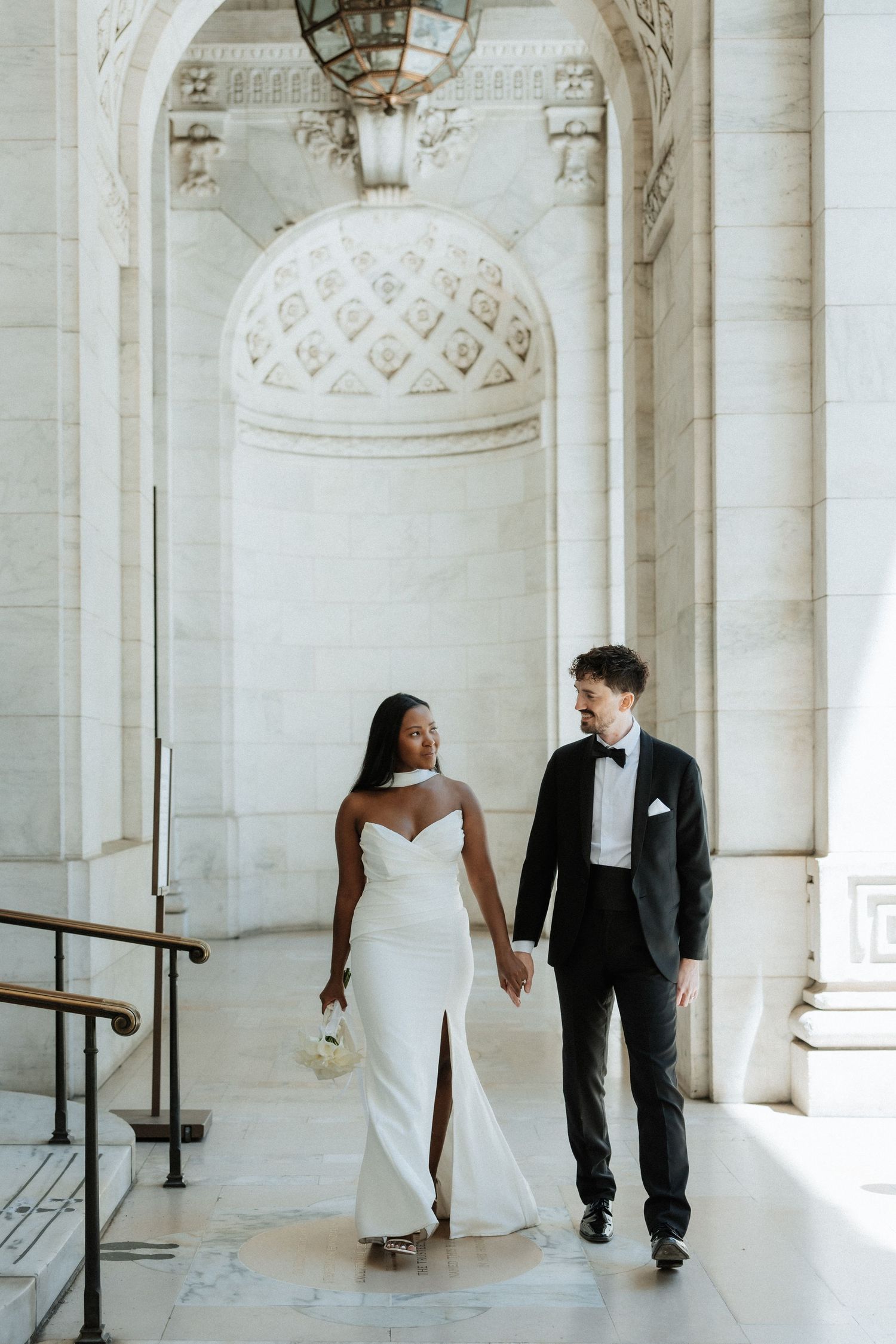 A couple poses in wedding attire under an ornate white stone colonnade with detailed ceiling work.
