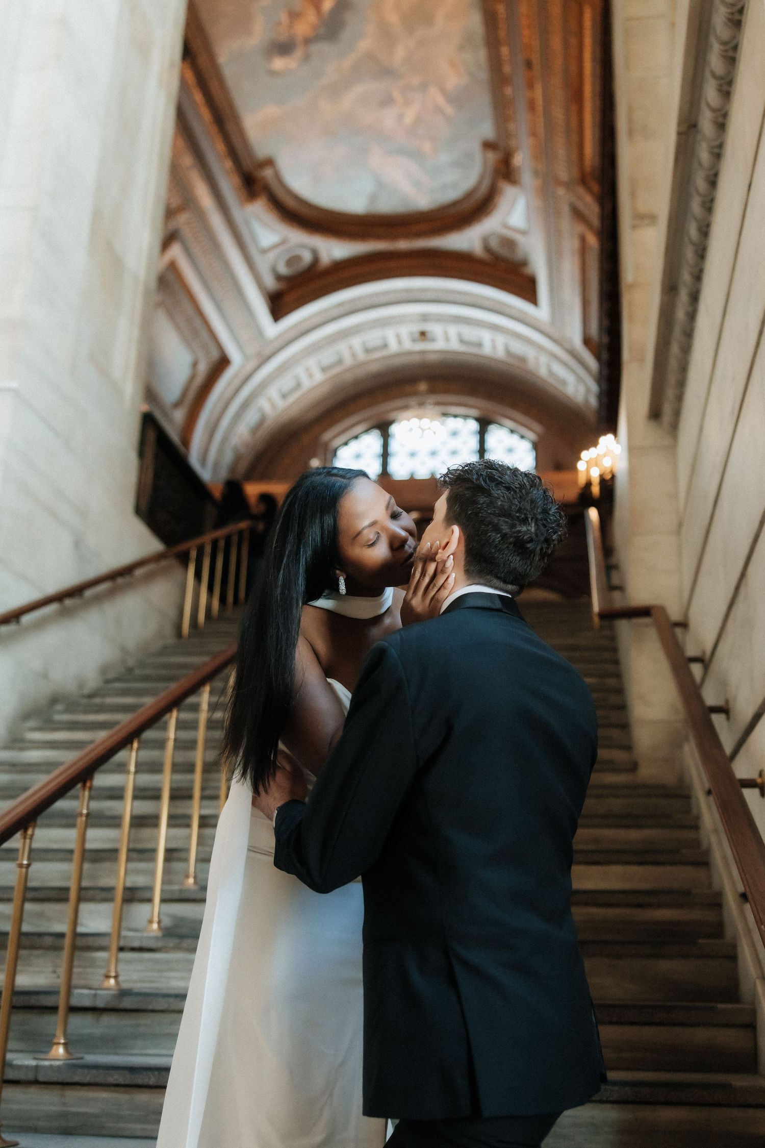 Romantic couple embraces on an elegant marble staircase under ornate architecture and a fresco ceiling.