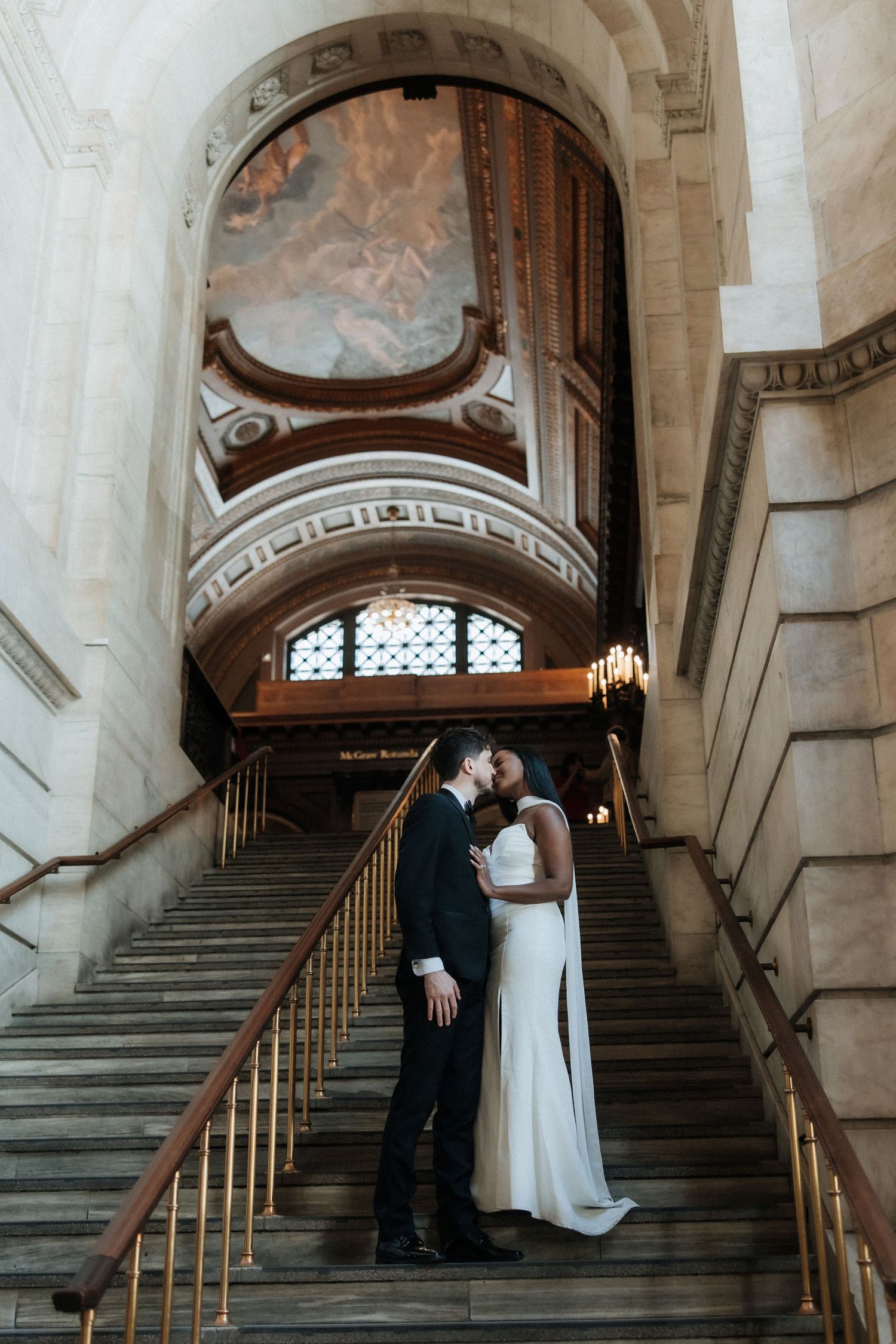 Wedding couple poses on a grand staircase with brass railings and painted archways.